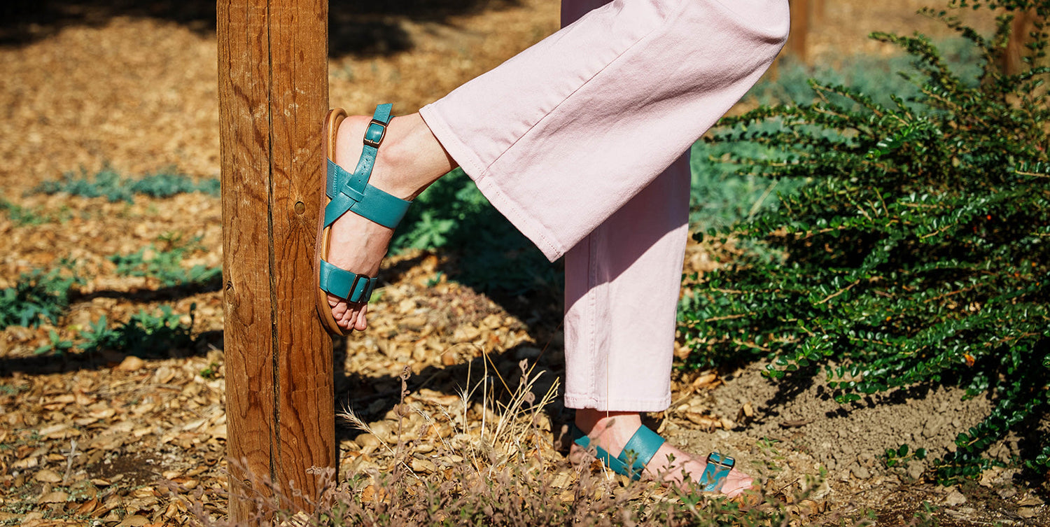 woman in blue jeans wearing triple groundz sandals lifting one leg on a metal garden chair